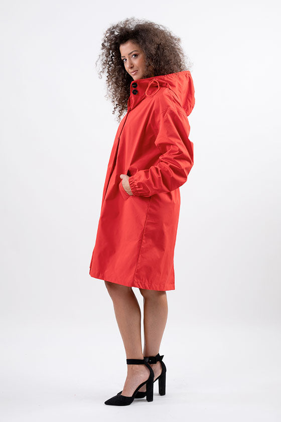 A person with curly hair is standing sideways, wearing a stylish CRIMSON RED WINDBREAKER by looselyboho that extends to their knees. They have their hands in the pockets and are wearing black high-heeled shoes. The background is a plain white.